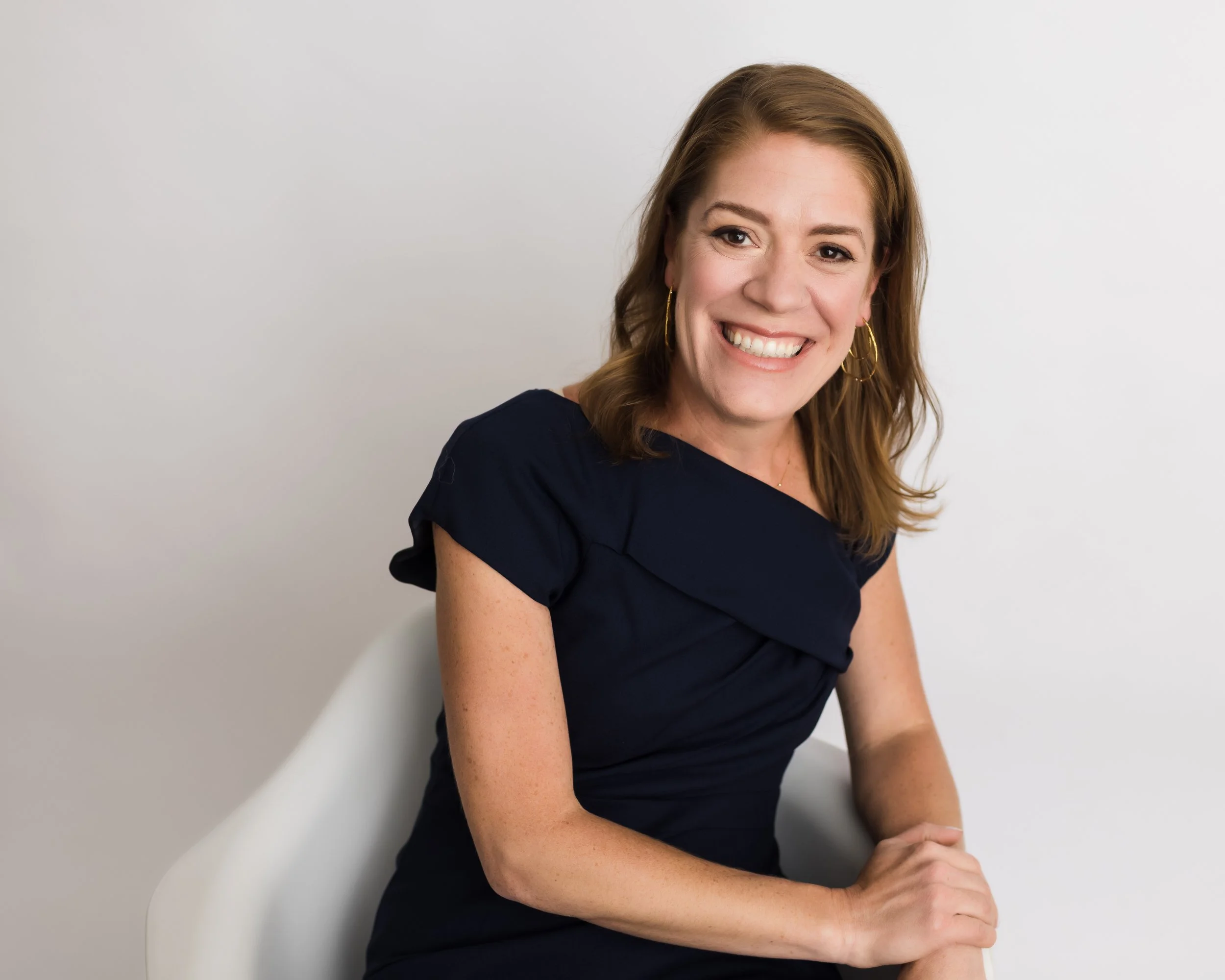 A woman with shoulder-length light brown hair, wearing a navy blue dress and gold hoop earrings, smiling while sitting on a white chair against a plain white background.