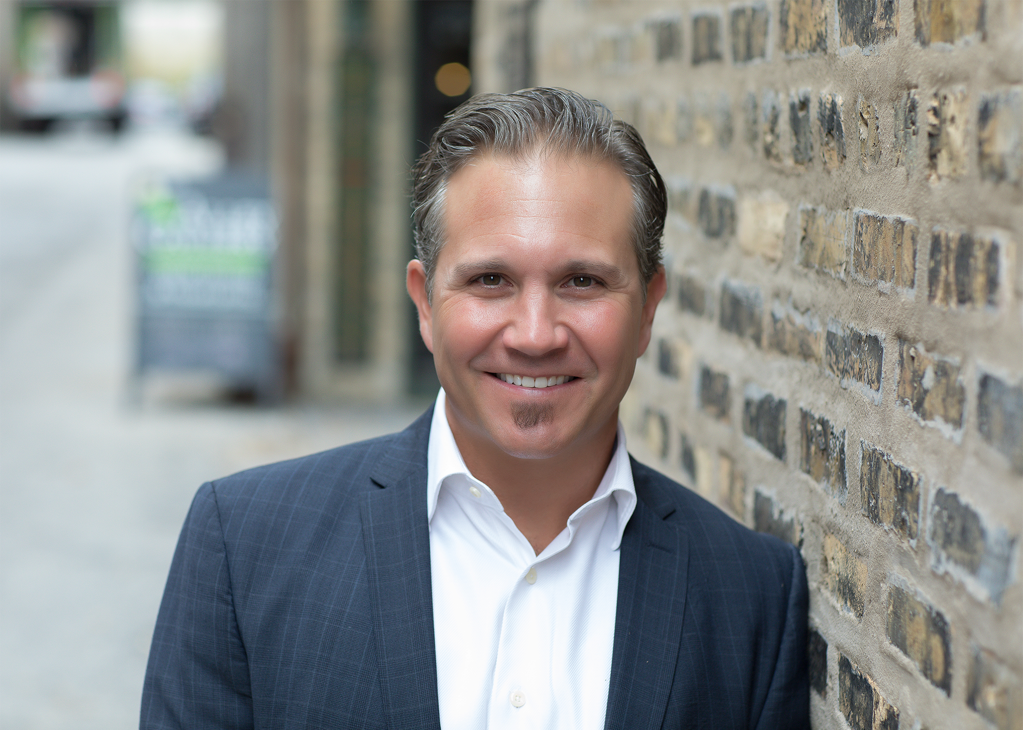 A professional man in a suit standing outdoors next to a brick wall, smiling at the camera.