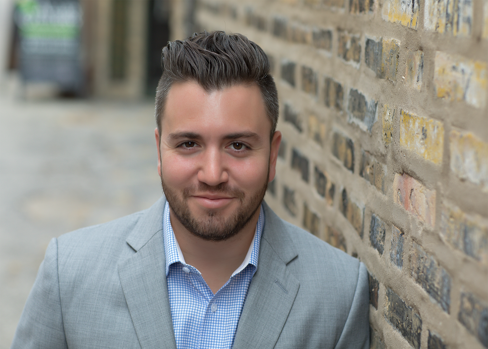 A man with dark hair and a beard wearing a light gray blazer and a blue checkered shirt, standing outdoors next to a brick wall.