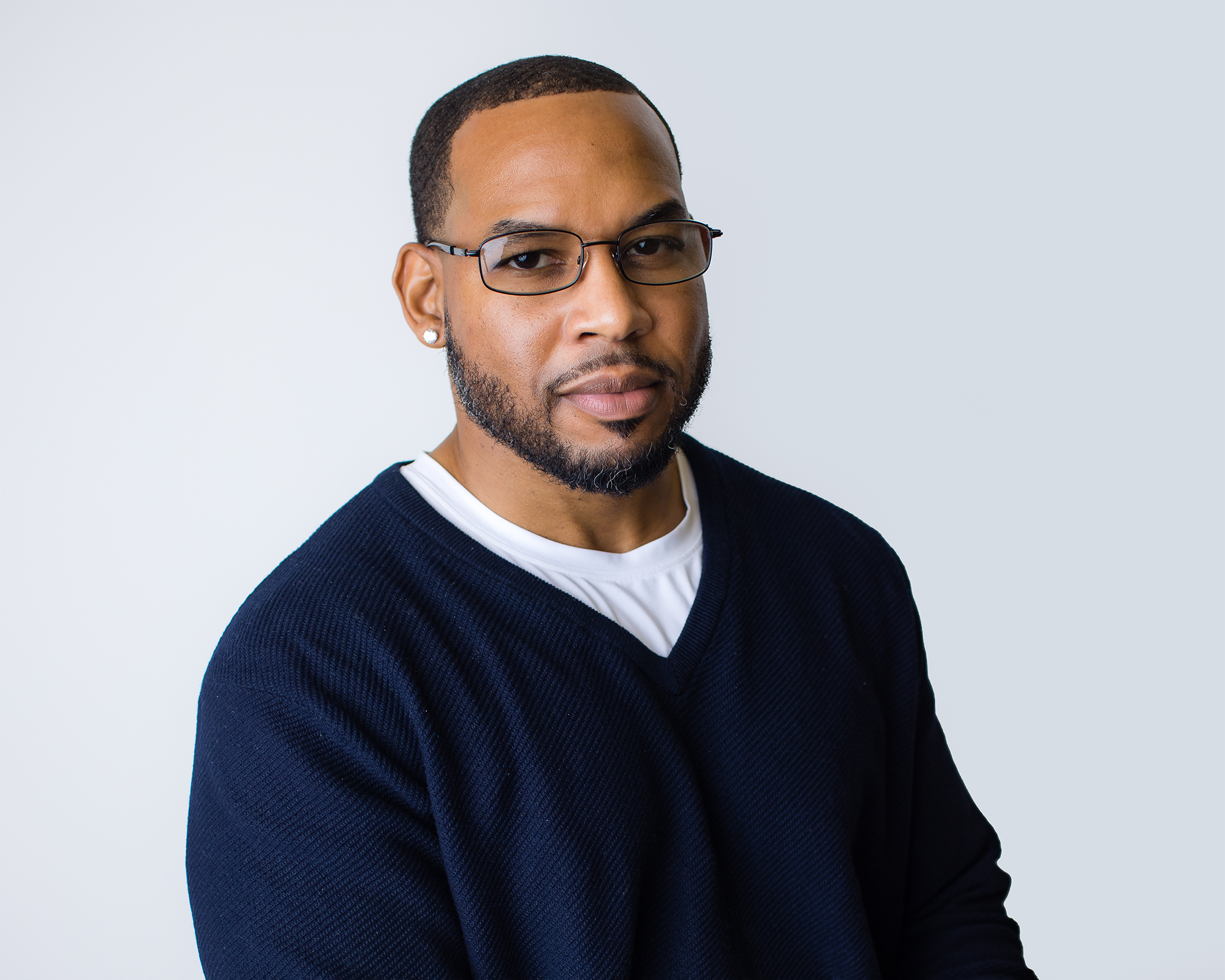 A man with short hair, wearing glasses, earrings, a white shirt, and a dark sweater, posing against a plain light background.