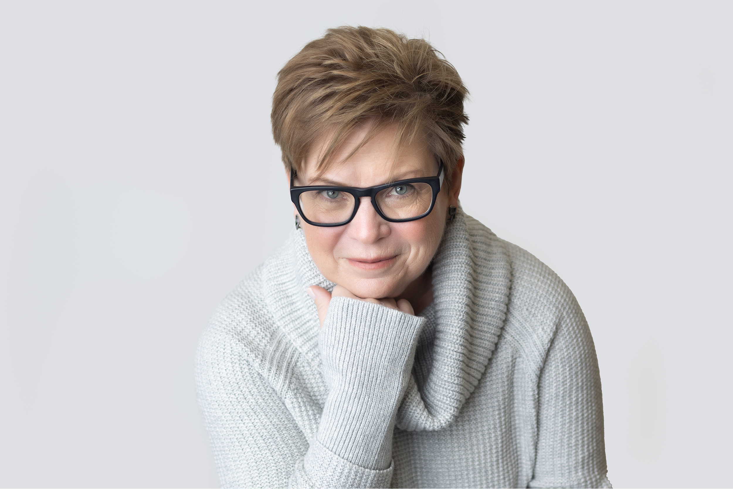 A middle-aged woman with short, light brown hair and glasses looking into the camera with a slight smile, resting her chin on her hand.
