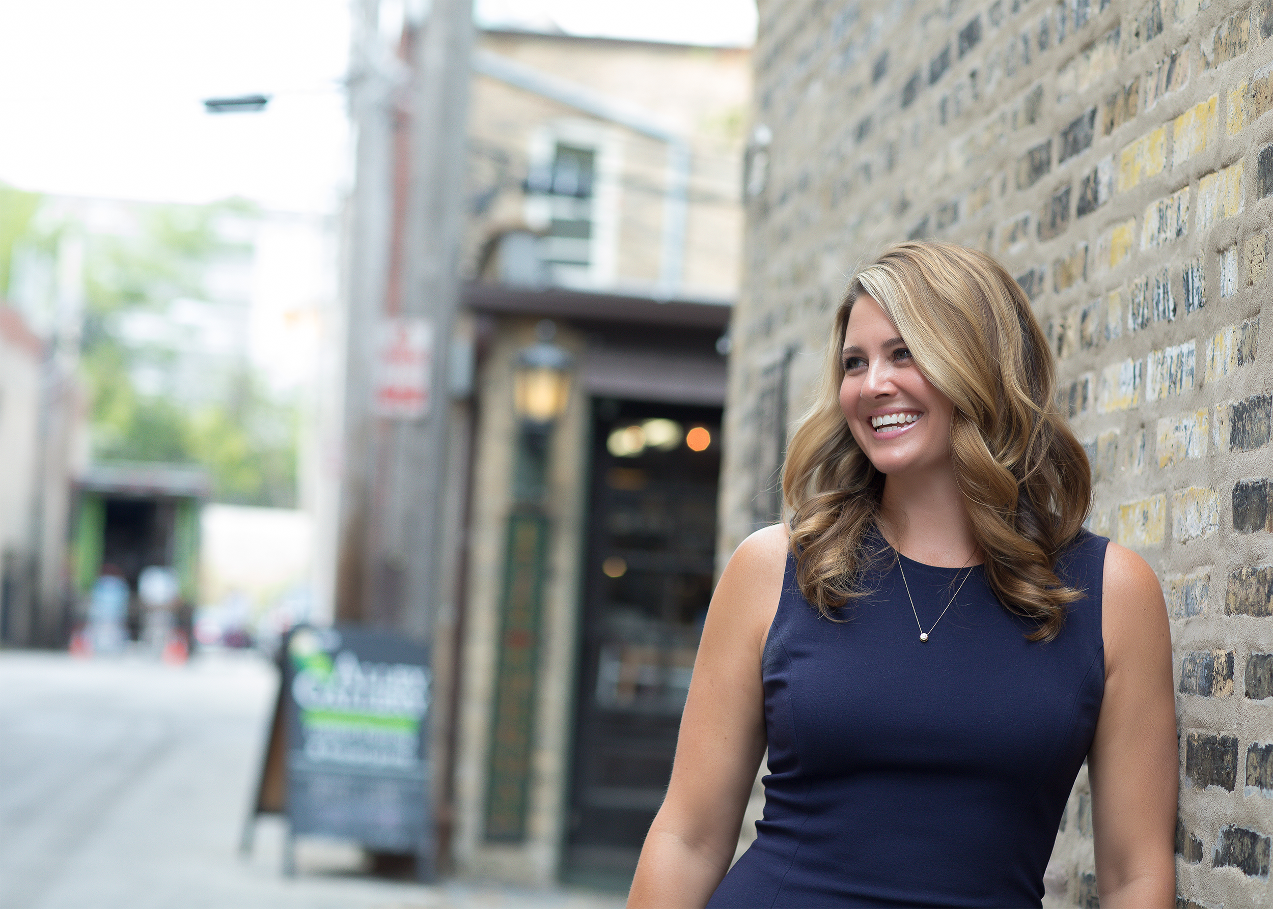 A woman with curly blonde hair wearing a navy sleeveless dress, smiling while leaning against a brick wall on an urban street.