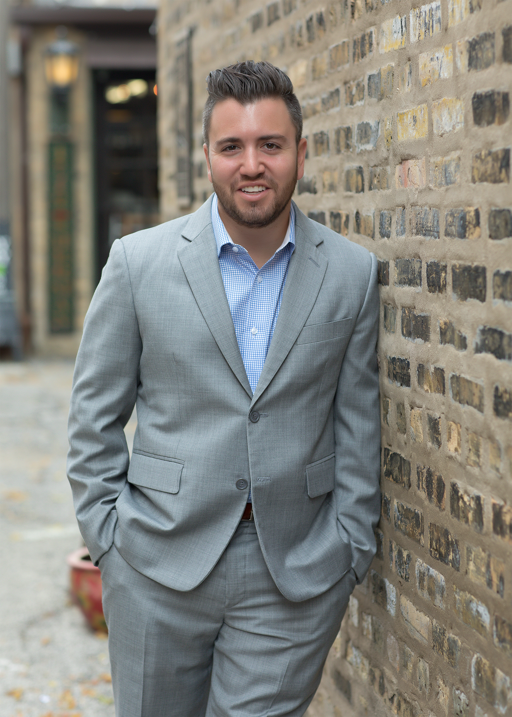 A man in a grey suit leaning against a brick wall outdoors.