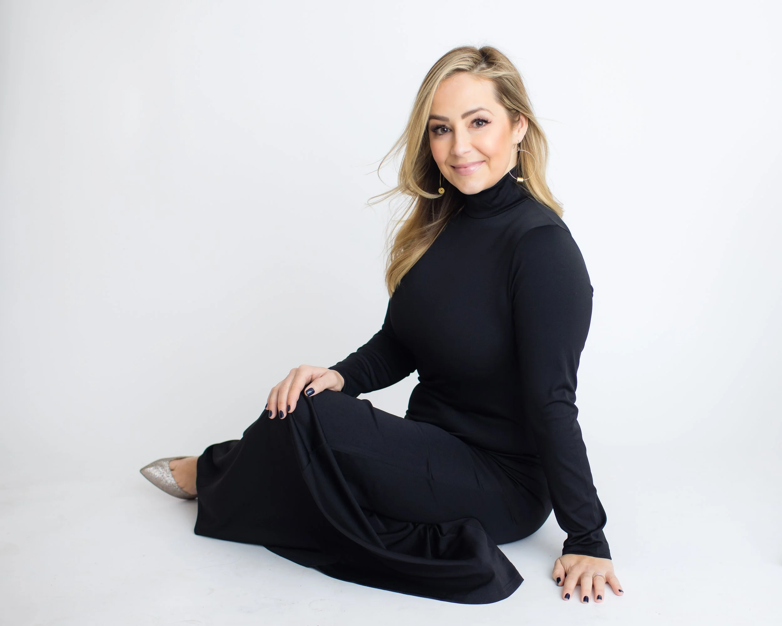 A woman sitting on the floor against a plain white background, wearing a black turtleneck and black pants, with blonde hair and makeup, smiling at the camera.