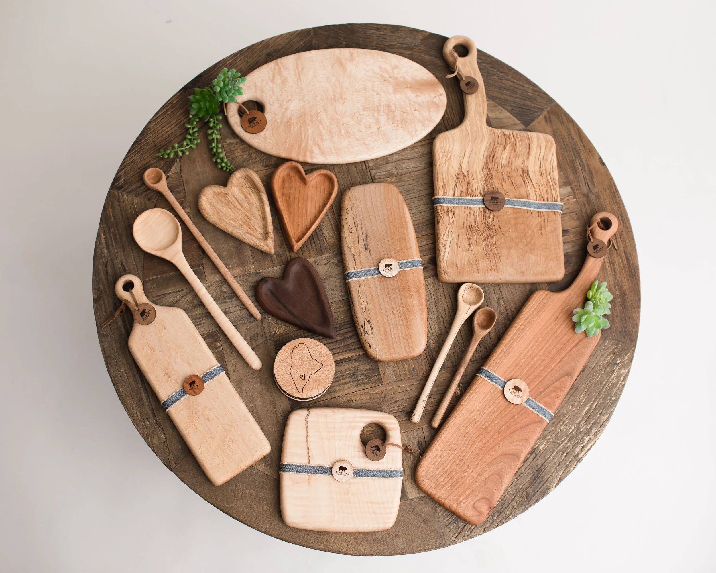 Various wooden kitchen utensils and decor items on a round wooden table, including cutting boards, spoons, hearts, a small plate, and succulent plants.