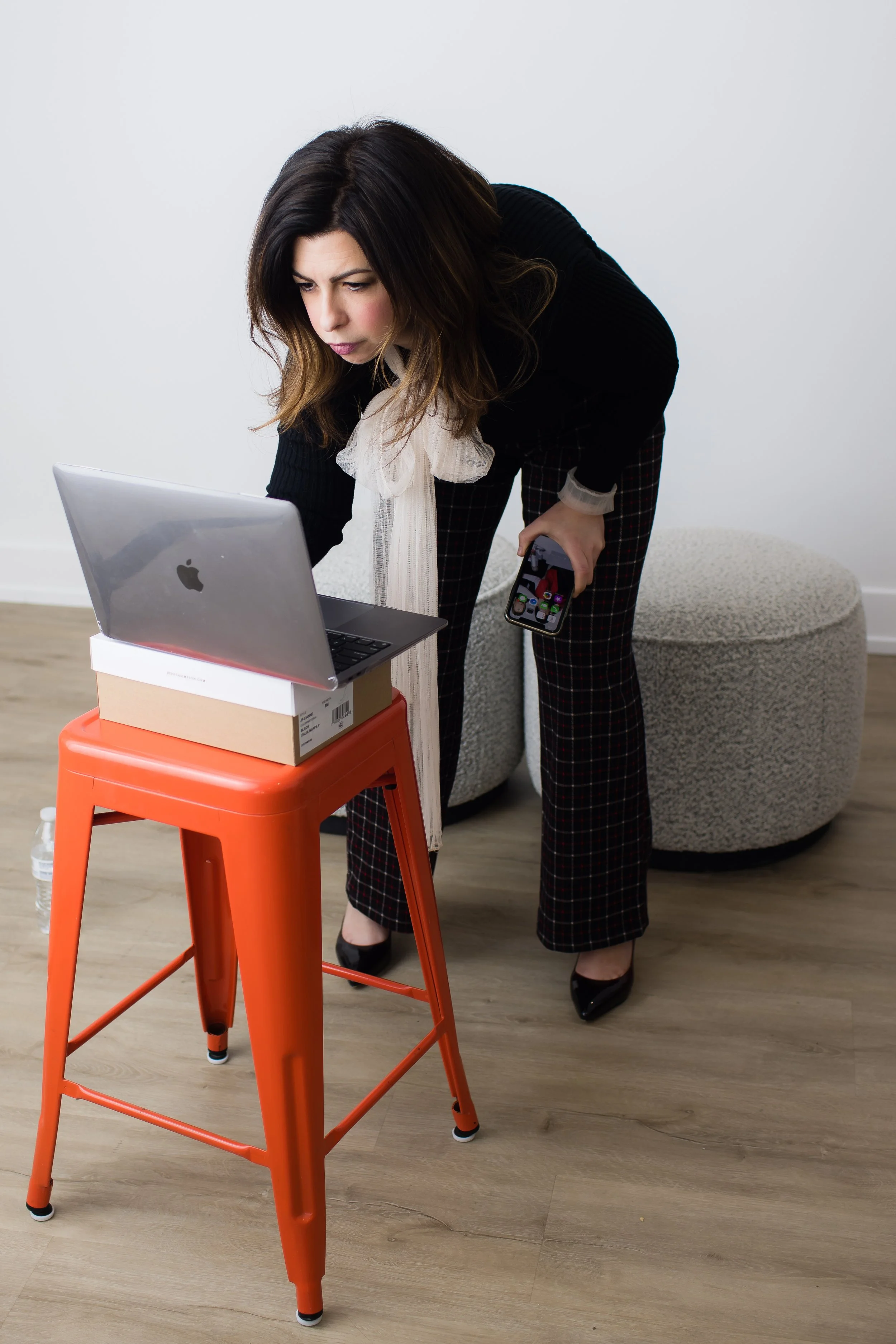 Woman leaning over on a stool, looking at a laptop and holding a phone in her hand in a room with a gray ottoman and wooden floor.