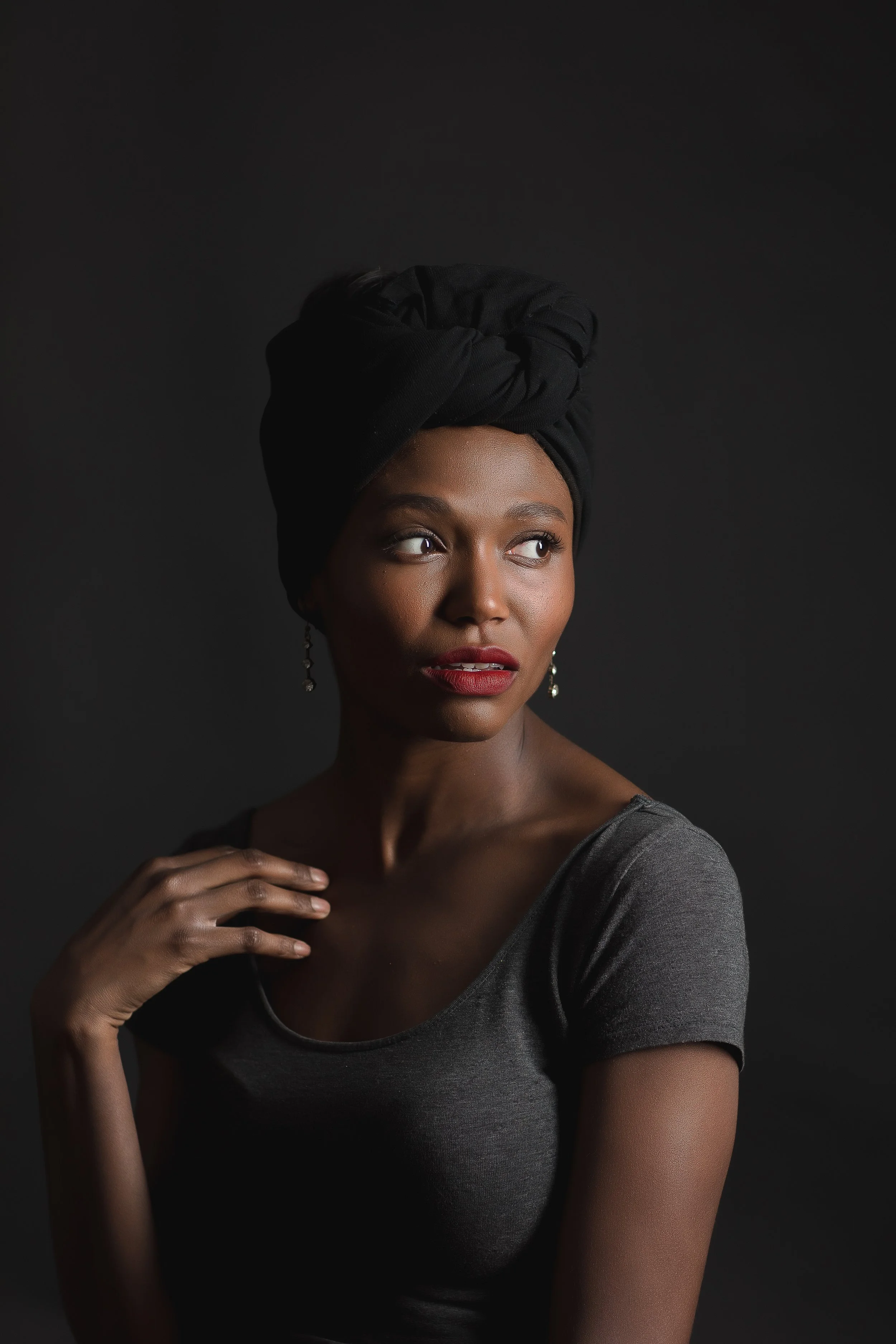 A woman with dark skin wearing a black headwrap and gray top, looking to her right against a dark background.