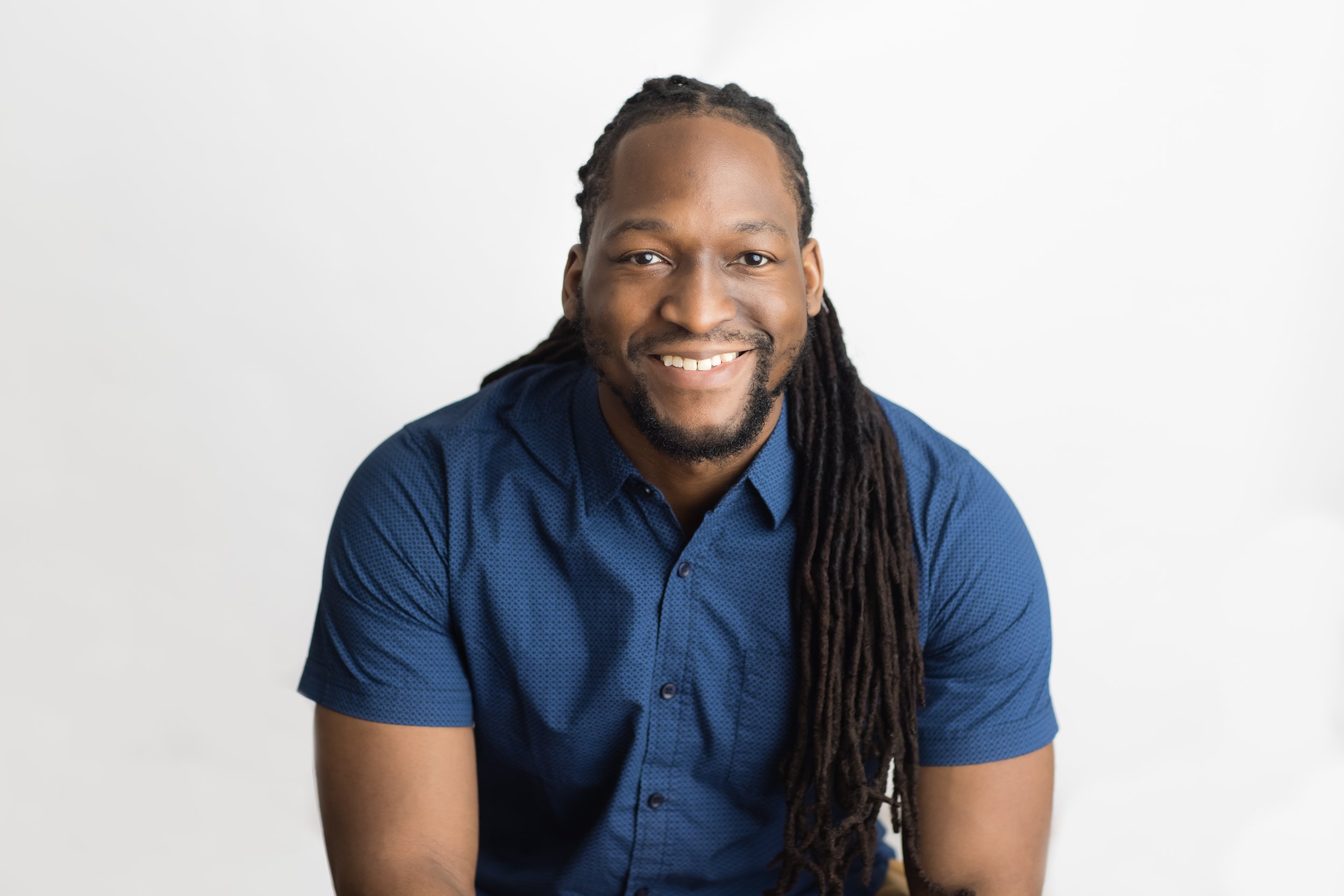 Smiling man with long dreadlocks wearing a blue button-up shirt against a white background.