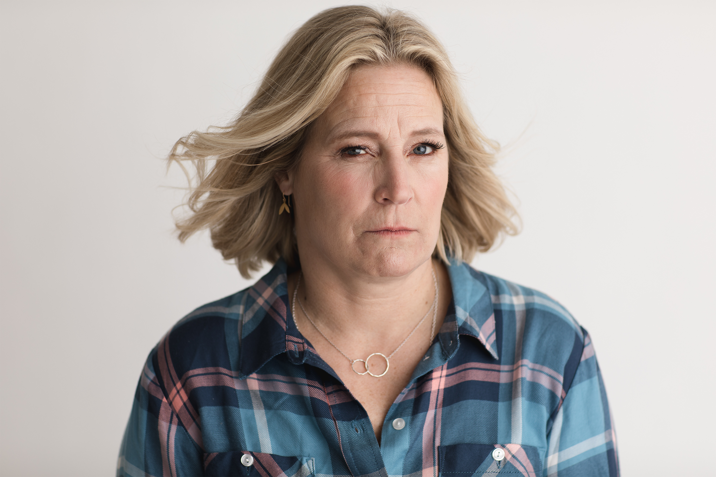 A middle-aged woman with blonde hair styled in loose waves, wearing a blue plaid shirt and silver jewelry, looking with a serious expression against a plain white background.