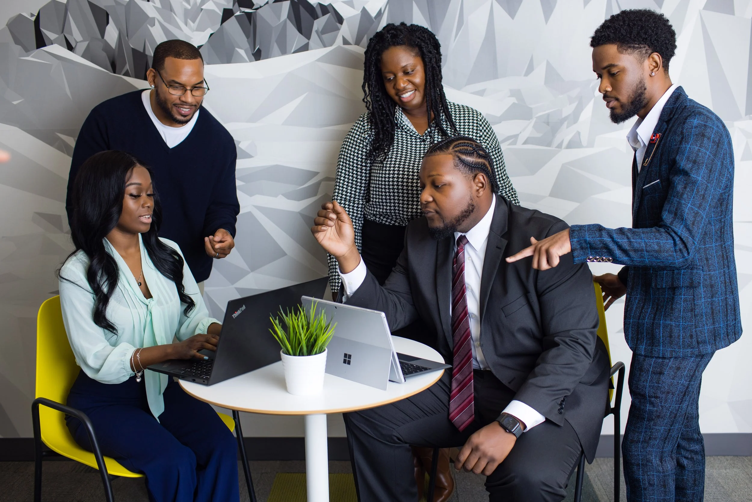 Five diverse business professionals in an office around a table, engaged in a discussion and collaboration, with laptops and a plant on the table.