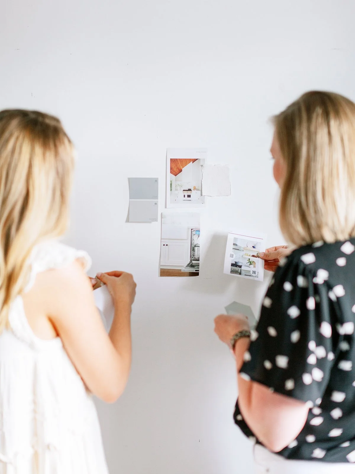 Two women discussing interior design ideas with mood boards and photos on a white wall.