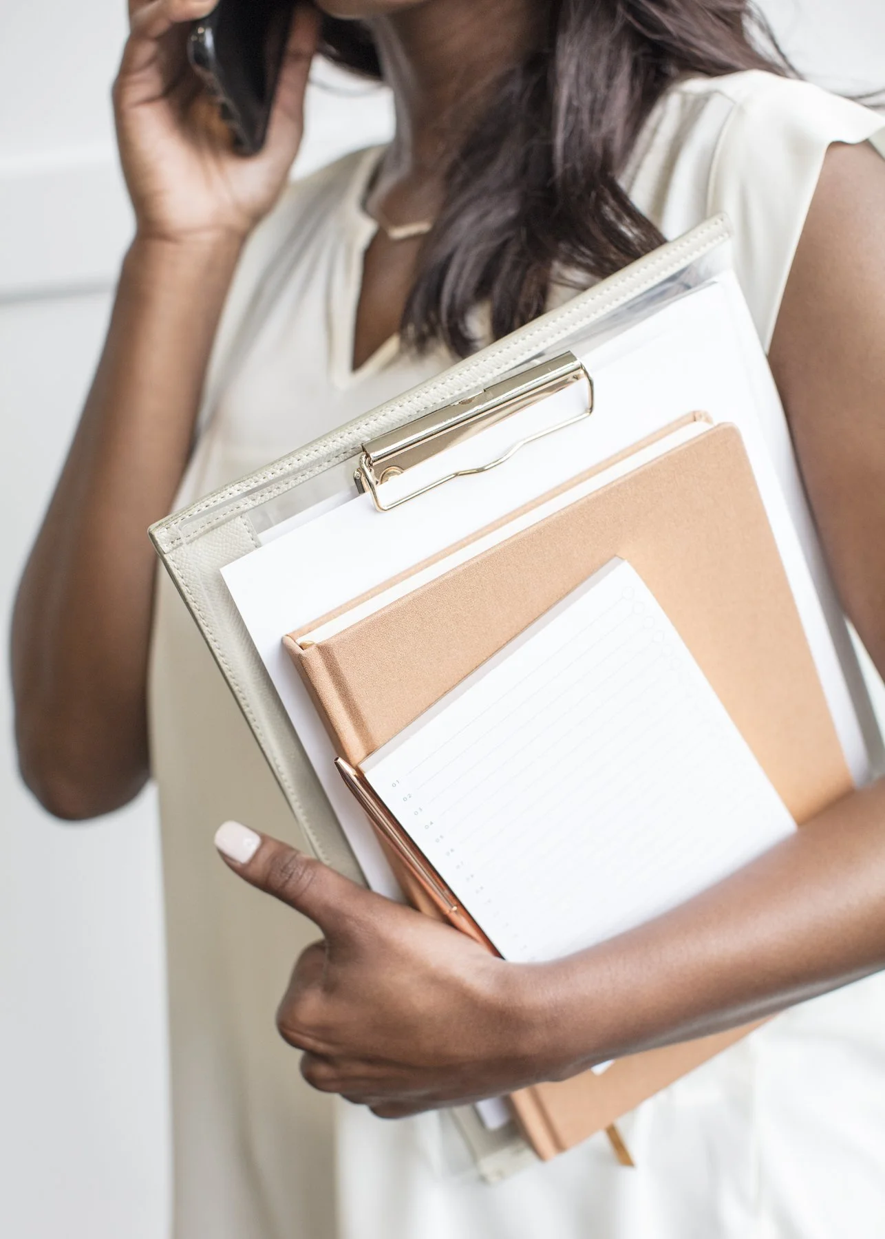 A woman holding a clipboard with yellow and white notebooks, a sheet of paper, and a orange pen, while talking on a cell phone.