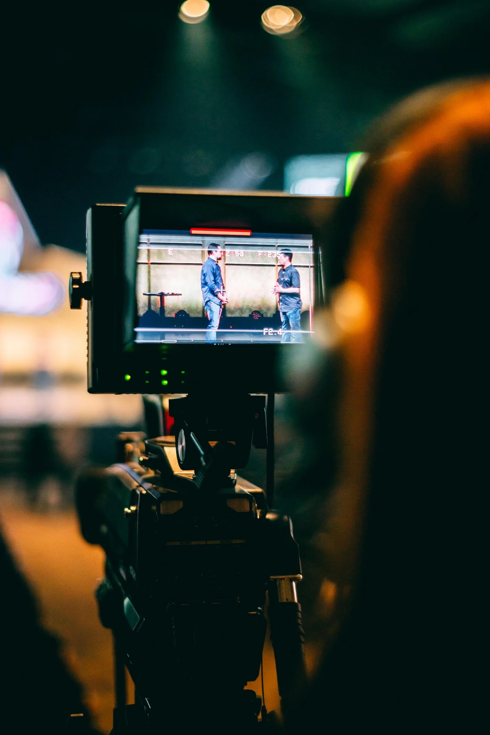 A camera screen capturing two people standing and talking to each other, with a blurred background of a stage or studio environment.