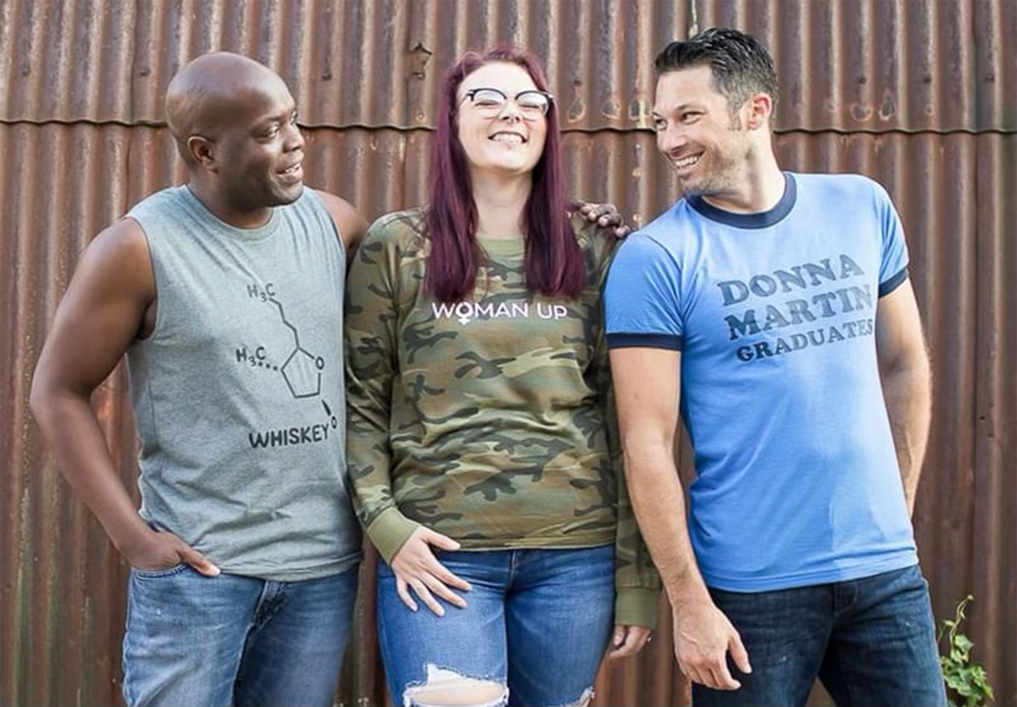 Three friends standing together outdoors, smiling and looking at each other, with rusted metal wall in the background. The woman in the middle is wearing a camo shirt that says 'Woman Up'. The man on the left is wearing a sleeveless gray shirt with a whiskey chemical structure. The man on the right is wearing a blue T-shirt that says 'Donna Martin Graduates'.