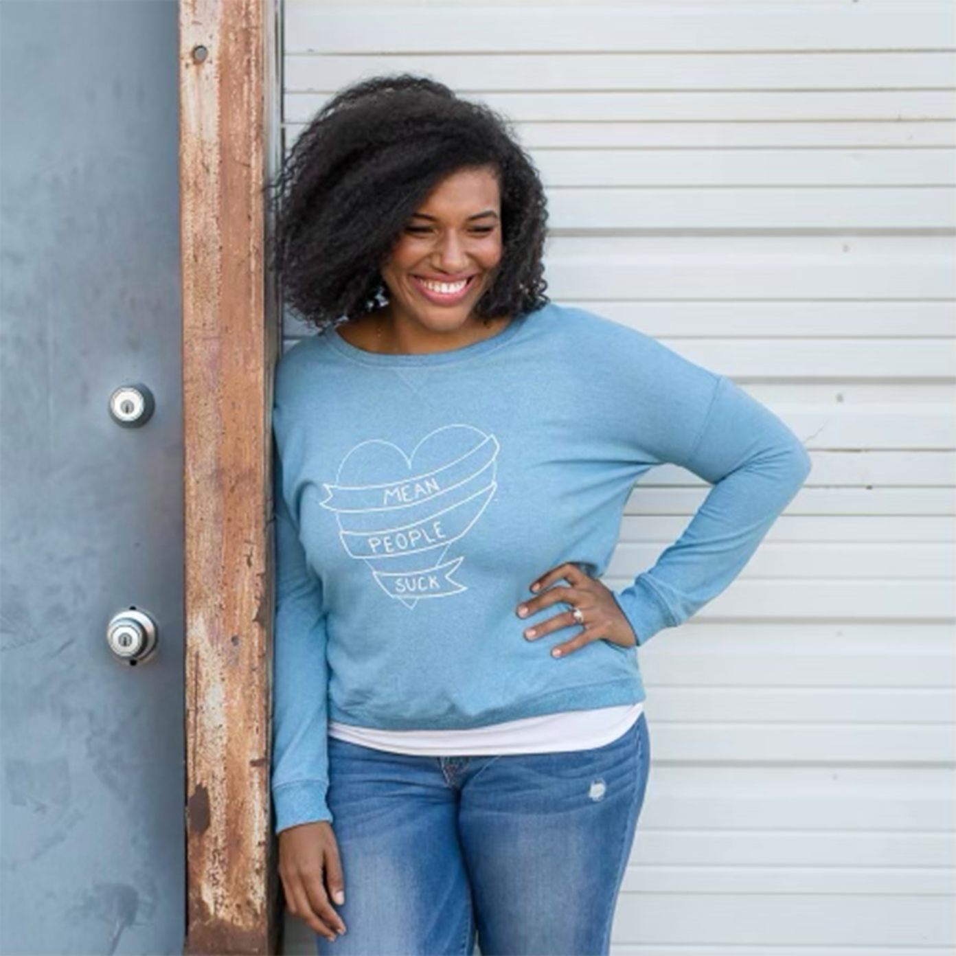 A woman with curly hair smiling, wearing a blue sweatshirt with the words "Mean People Suck" on it, standing next to a door with her hand on her hip.