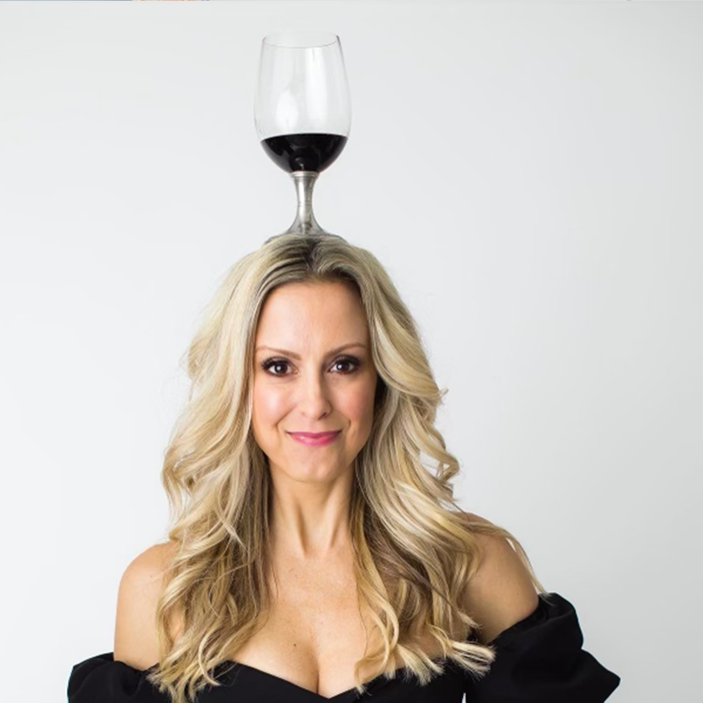 A woman with long blonde hair and a black off-the-shoulder top has a glass of red wine balanced on her head.