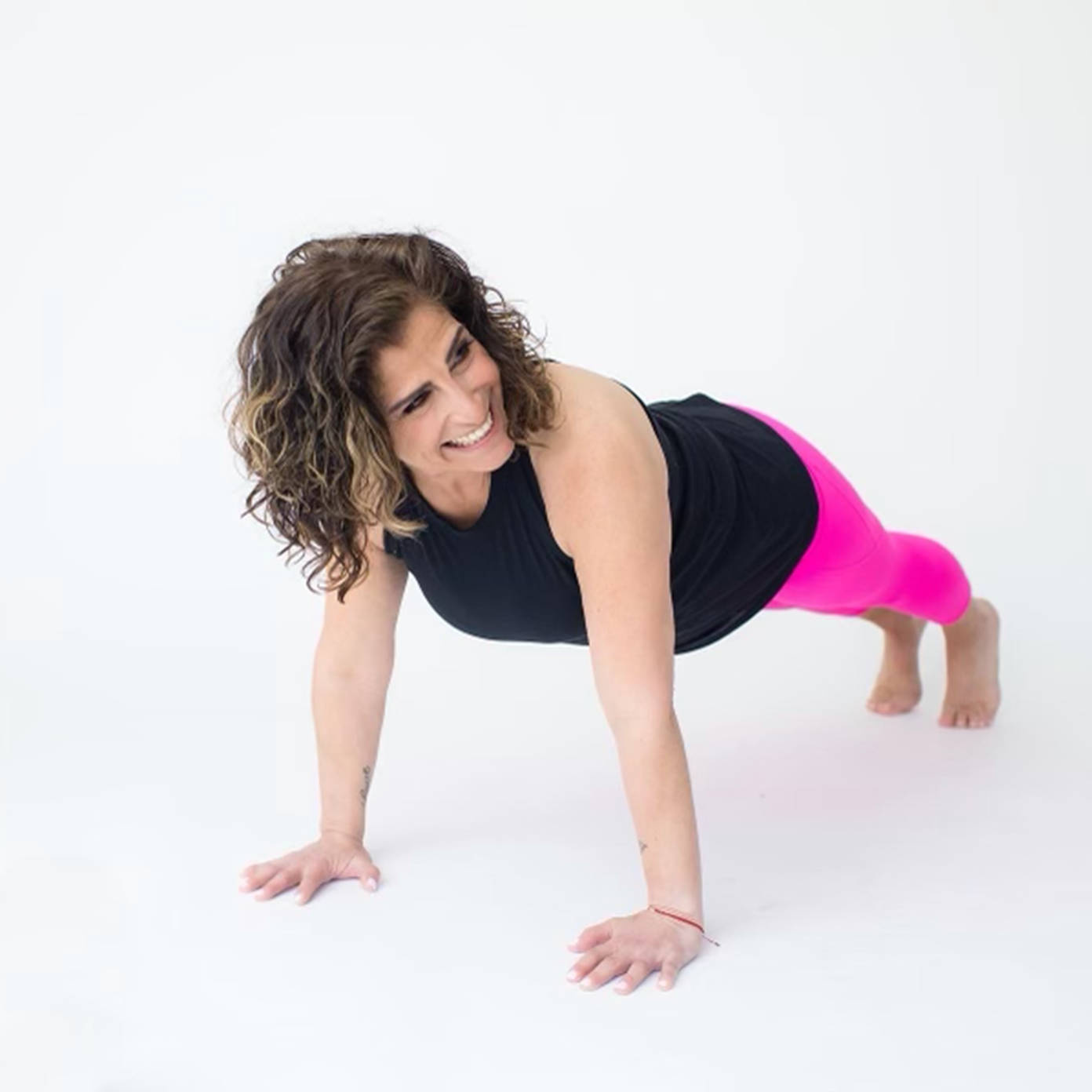 Woman doing a plank exercise on a white background, wearing a black top and pink leggings, smiling.