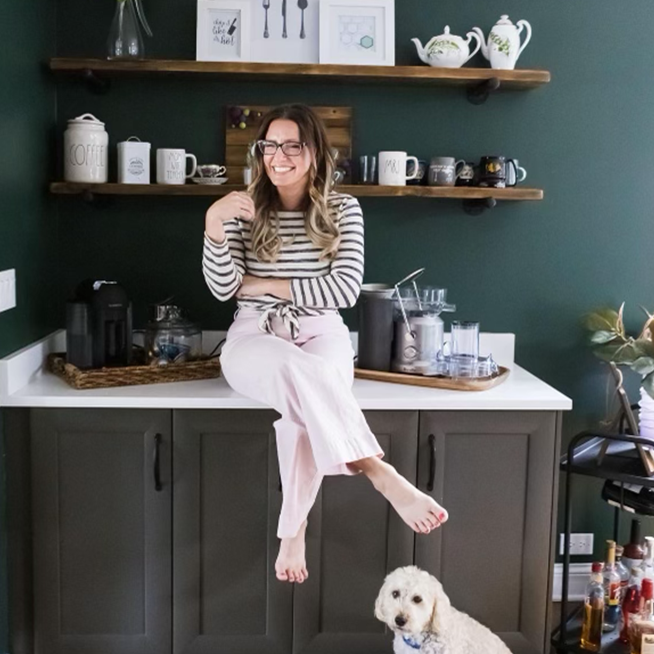 A woman with long, wavy hair wearing glasses, a striped top, and pale pink pants sitting on a kitchen counter with a small puppy underneath, smiling and looking at the camera in a modern kitchen with dark cabinets, wooden shelves, and various kitchenware.