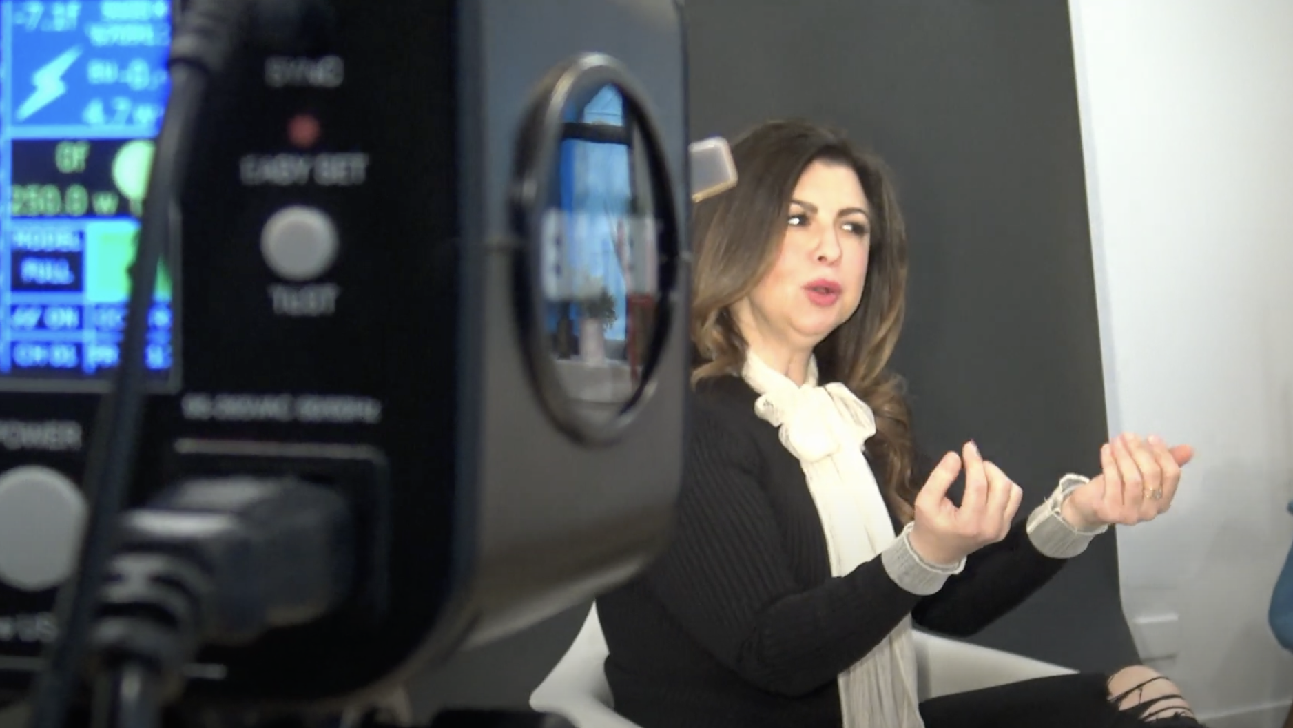 A woman with brown hair talking and gesturing with her hands during an interview, with professional camera equipment in the foreground.
