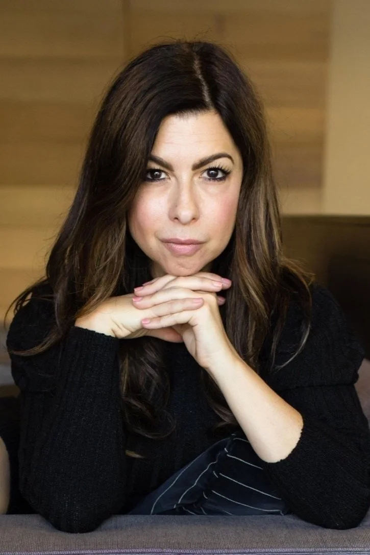 A woman with long brown hair, wearing a black sweater, resting her chin on her hands, looking directly at the camera in a cozy, warmly lit room.
