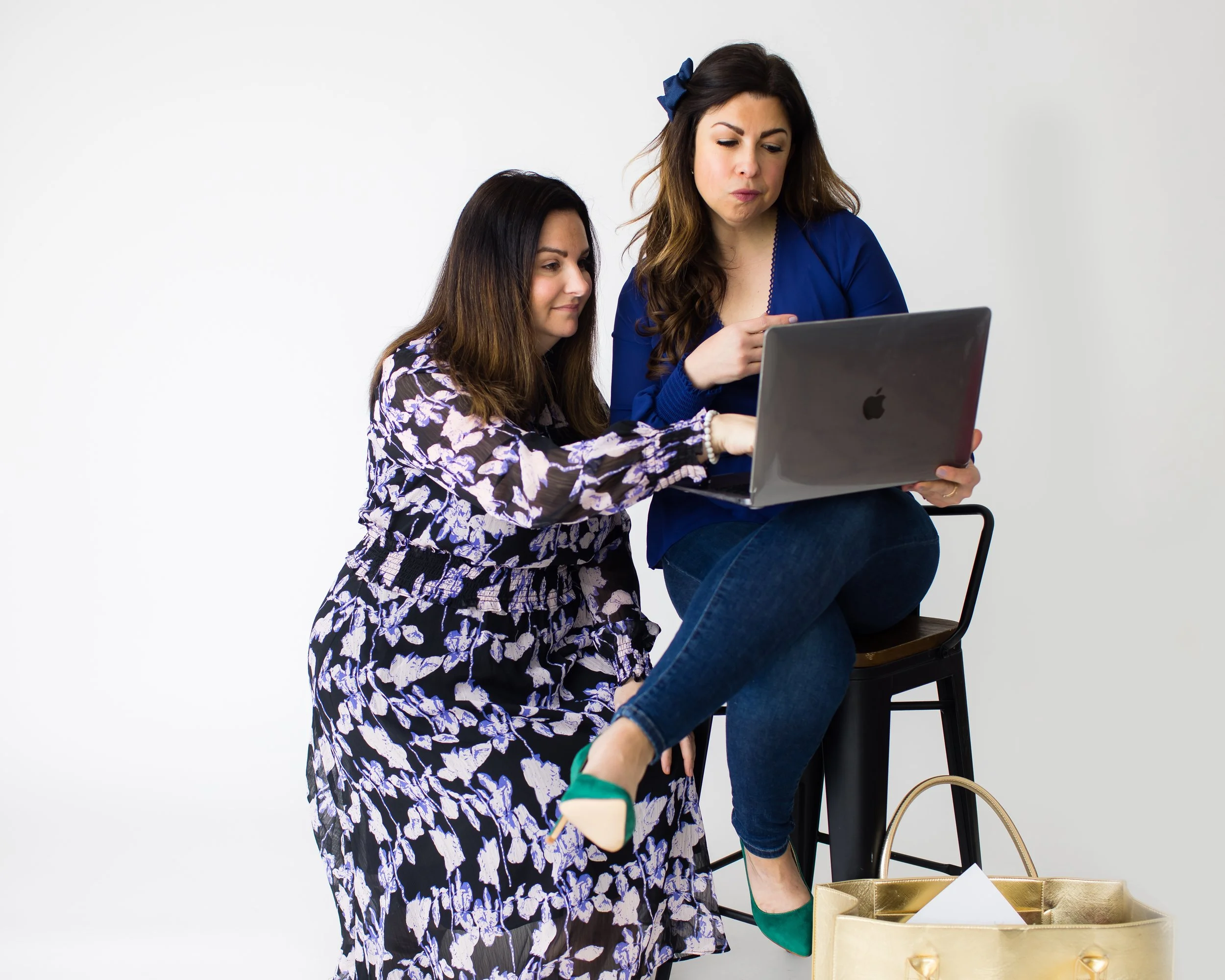 Two women looking at a laptop, one sitting on a stool and the other kneeling beside her, against a white background. The seated woman wears a blue top and jeans, and the kneeling woman wears a floral dress. There is a large gold handbag on the floor.