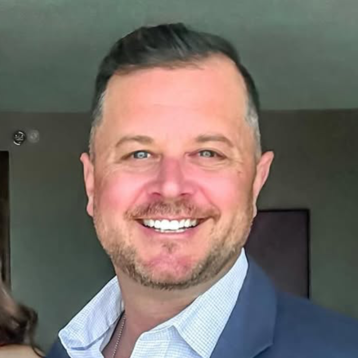 Smiling man in a suit with a light blue shirt and dark blue blazer, indoors.