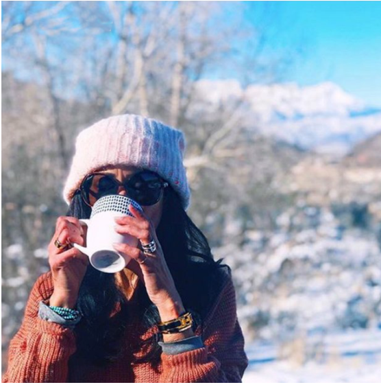 Woman wearing pink beanie and sunglasses drinking from a white mug outside in winter with snow-covered trees in the background.