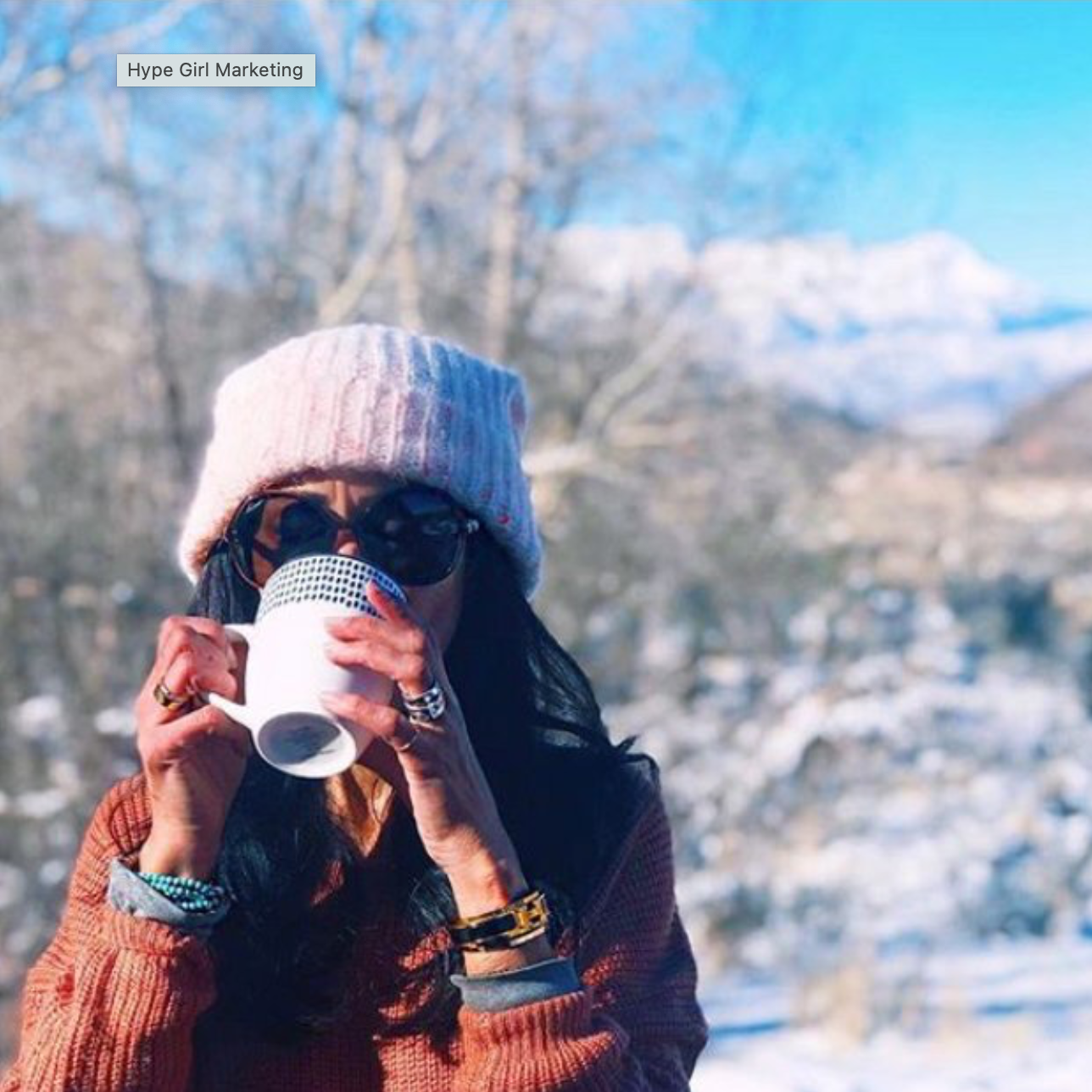 Woman in winter clothing, wearing a pink knit hat, sunglasses, drinking from a white mug outdoors in a snowy landscape with trees and mountains in the background.