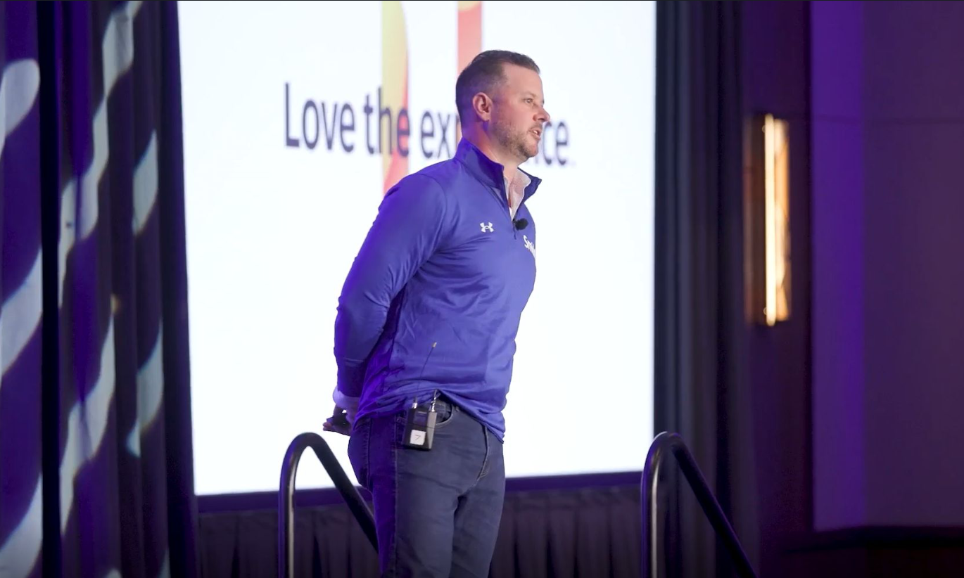 A man in a blue jacket on stage with a microphone clipped to his pocket, standing in front of a large screen displaying the partial message 'Love the ex'.