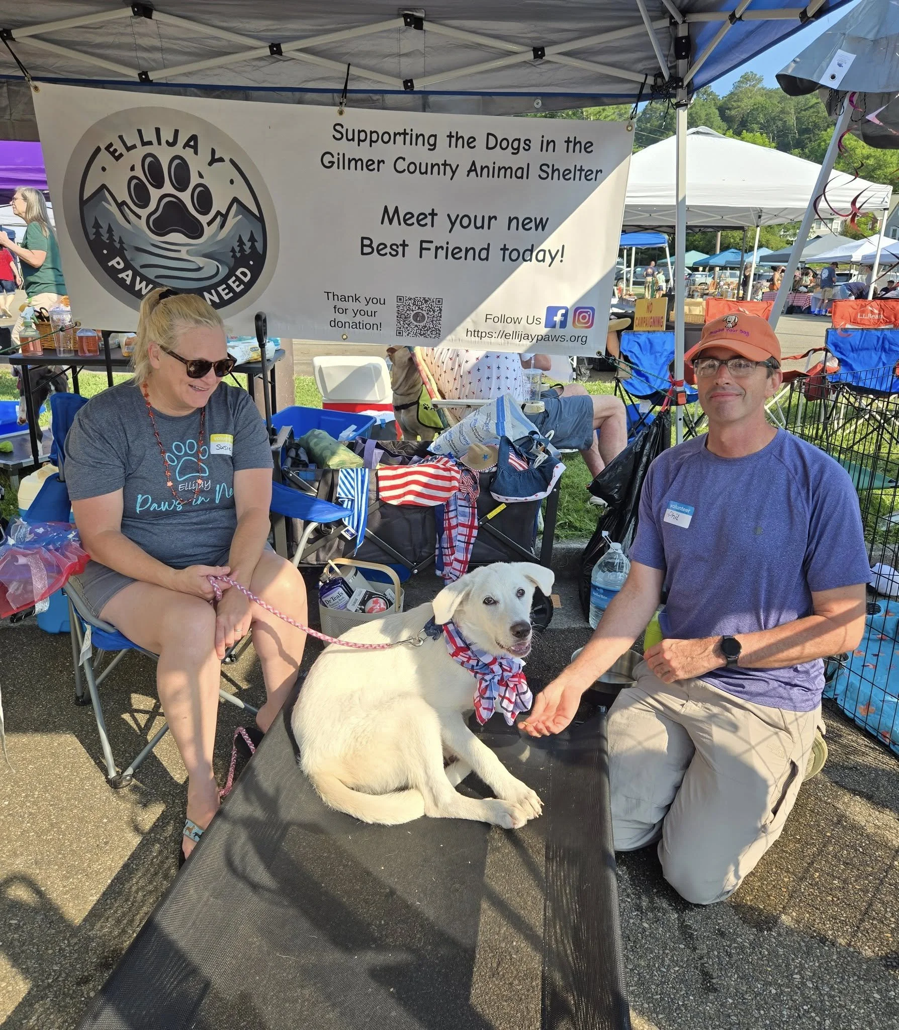 A man and woman at a pet adoption event with a white dog wearing a patriotic bandana. The woman is smiling and seated, the man kneels next to the dog, holding its paw. They are under a tent with a banner supporting Gilmer County Animal Shelter.