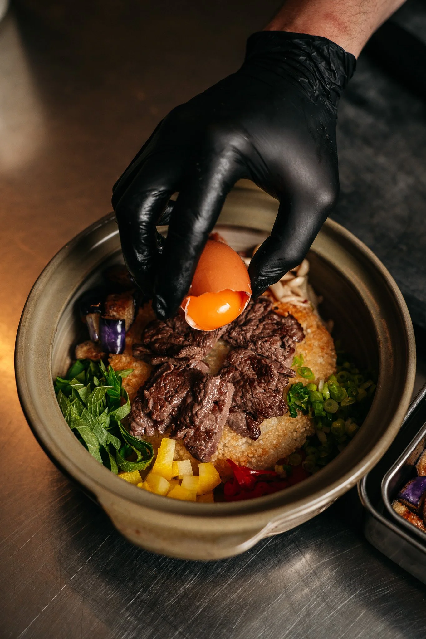 A hand wearing a black glove is placing a raw egg on top of a bowl of cooked ingredients including beef, vegetables, and rice.