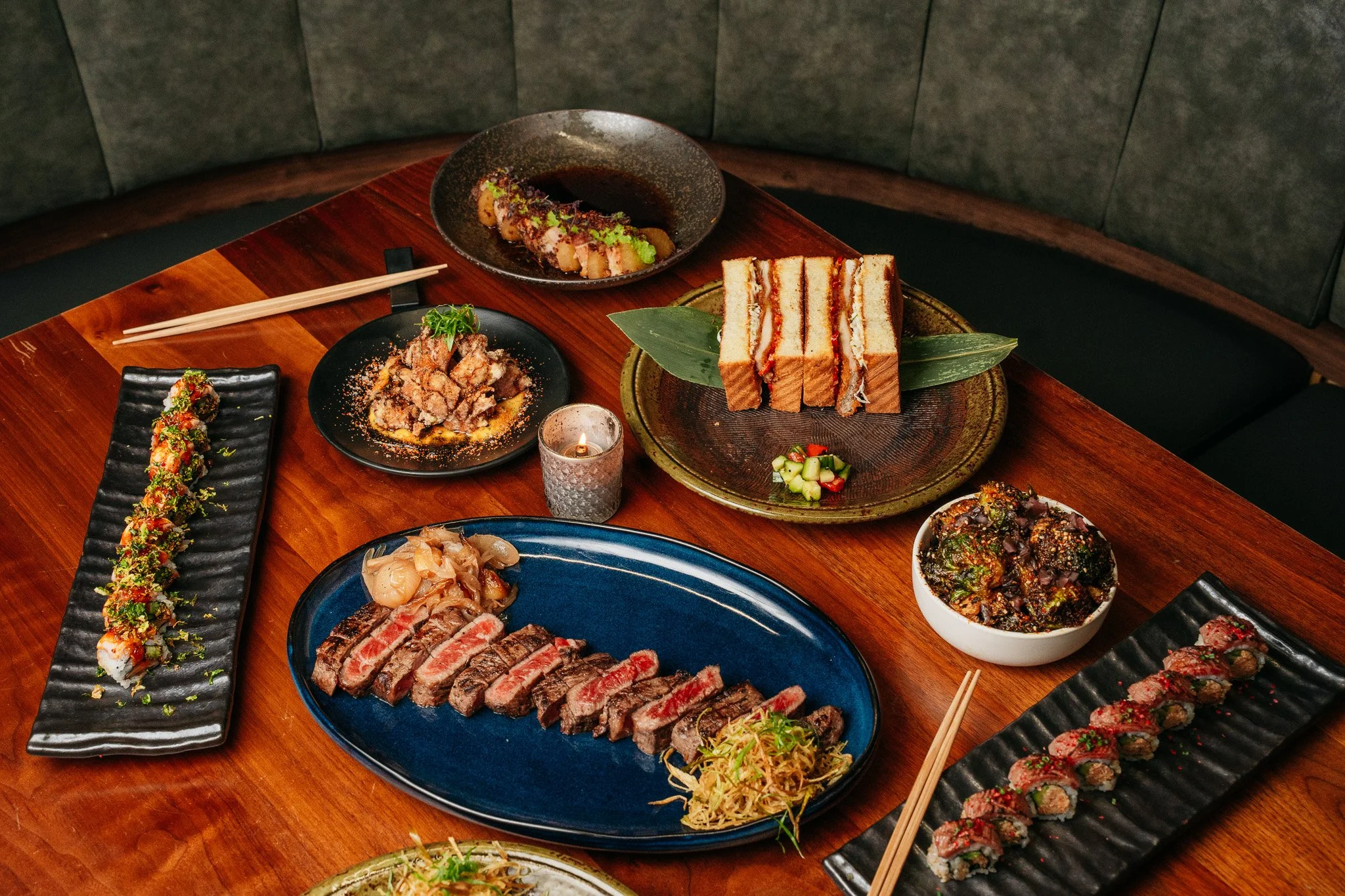 A variety of Japanese dishes, including sushi, sashimi, and other grilled or fried items, arranged on black, blue, and green plates on a wooden table.
