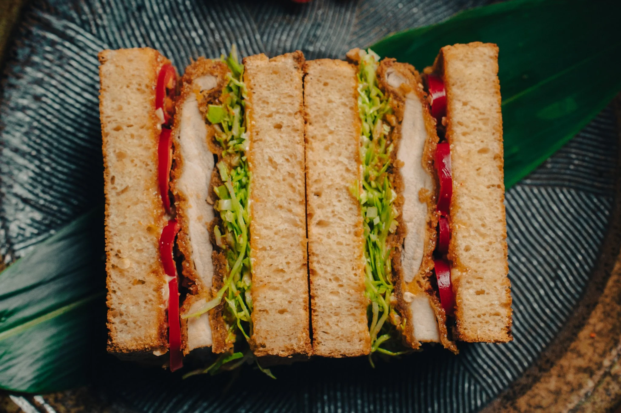 Triangular sandwich with crispy breaded chicken, lettuce, tomato, and mayonnaise on dark textured plate with green leaf underneath.