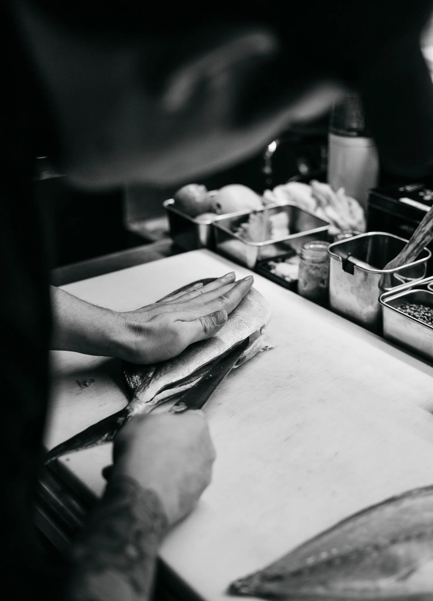 A person preparing a fish for cooking, with their hand resting on the fish on a cutting board, surrounded by kitchen utensils and ingredients.