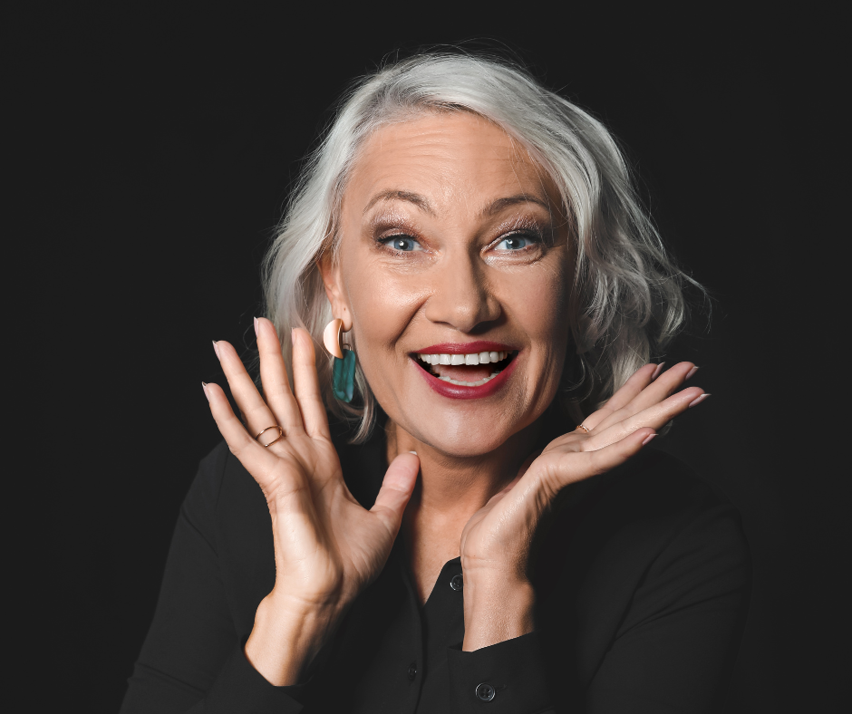 A joyful elderly woman with gray hair, wearing a black top, smiling widely and holding her hands near her face, set against a dark background.
