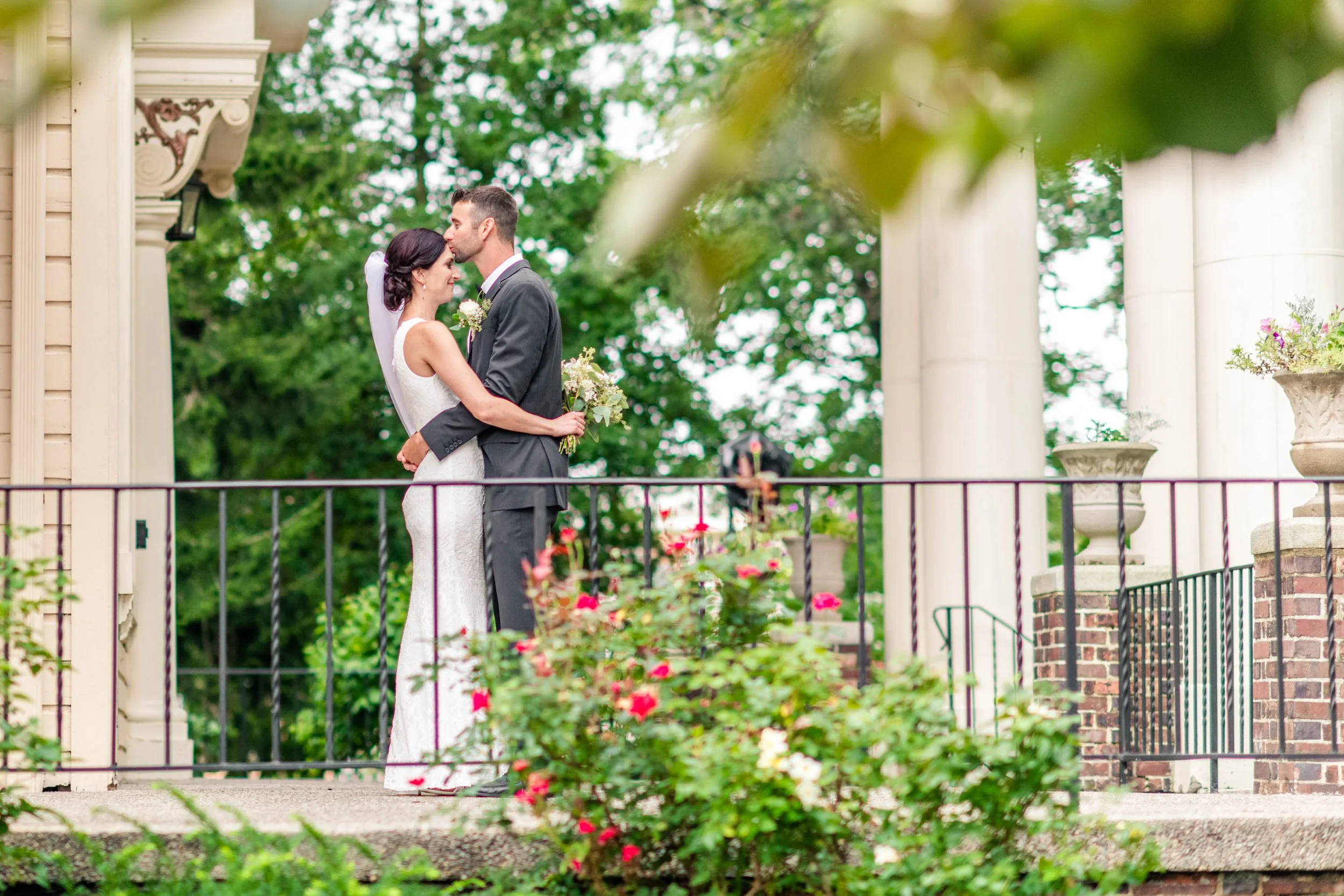 Bride and groom kissing on outdoor terrace with flowers and greenery, wearing wedding attire.