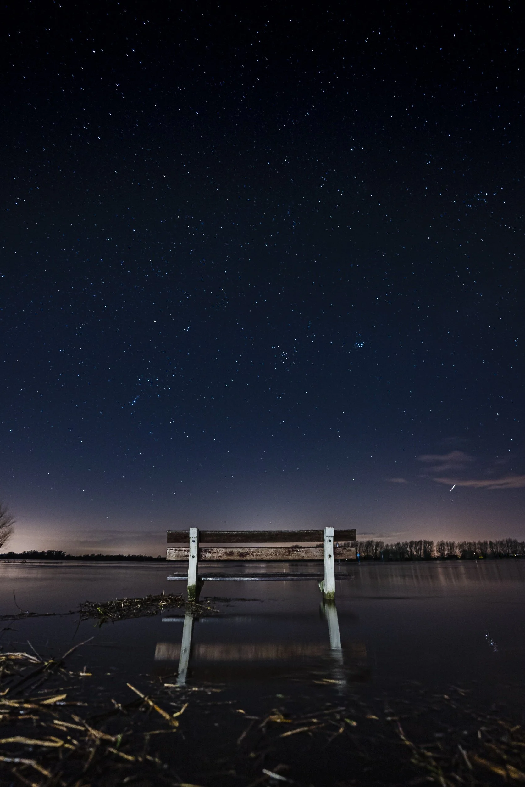 Houten bank onder sterrennacht aan de oever van een meer, met weerspiegeling in het water.