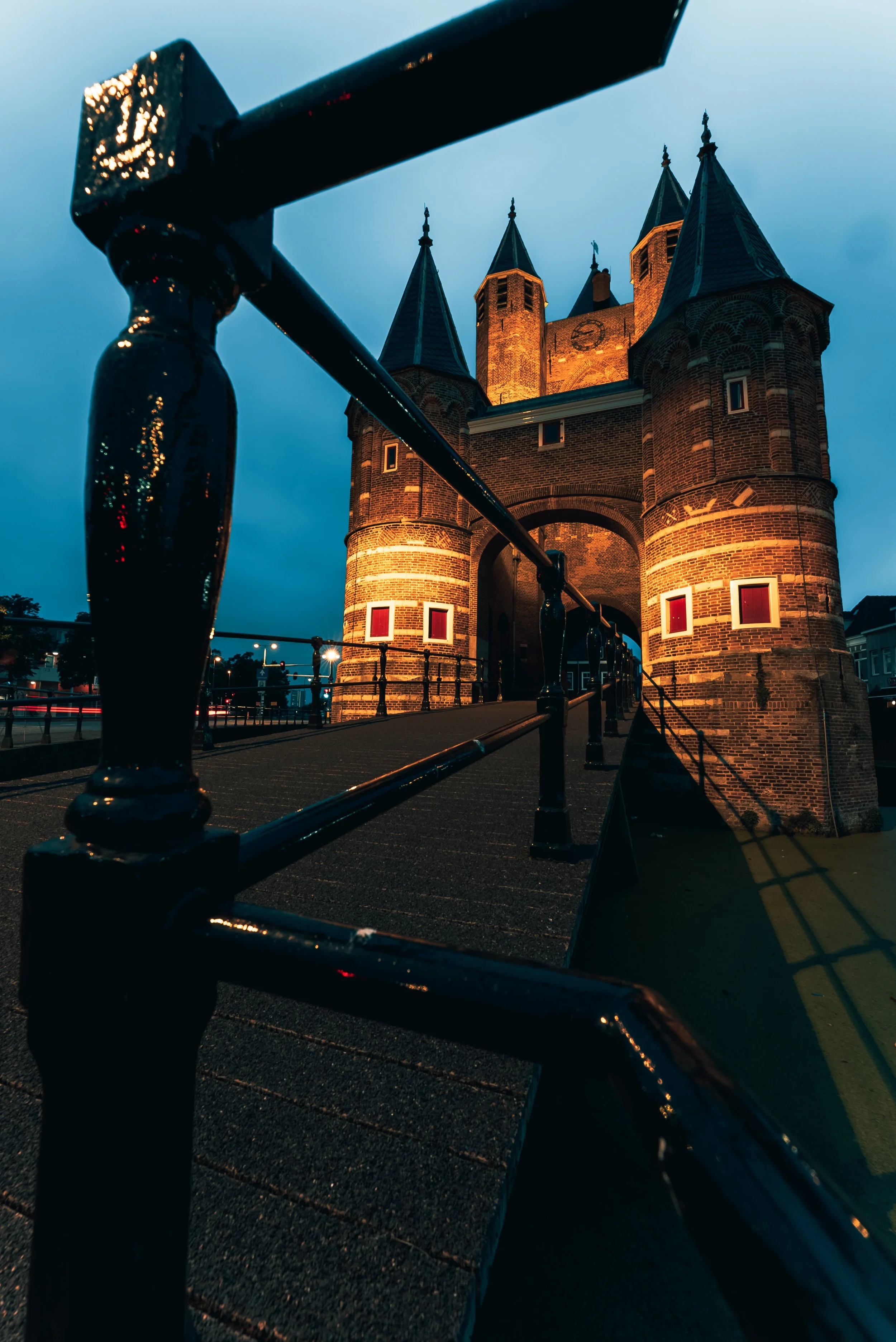 Avondfoto van een historische stenen poort met verlichte torens en een brug, gefotografeerd vanuit een laag hoekperspectief.