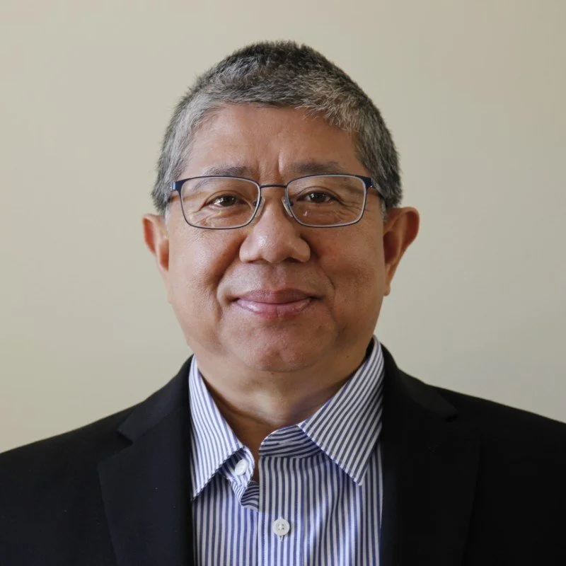 A middle-aged man with short gray hair, glasses, wearing a black blazer and a striped dress shirt, standing against a plain light-colored wall.