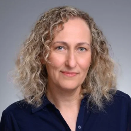 A woman with curly blonde hair wearing a navy blue collared shirt, posing against a neutral background.