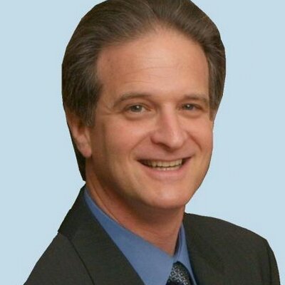 Headshot of a smiling man with dark hair in a suit and blue shirt against a light blue background.