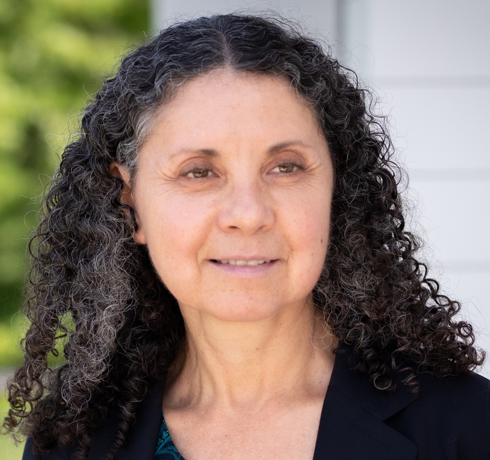 A woman with curly dark hair standing outside with a blurred background of trees and a white building.