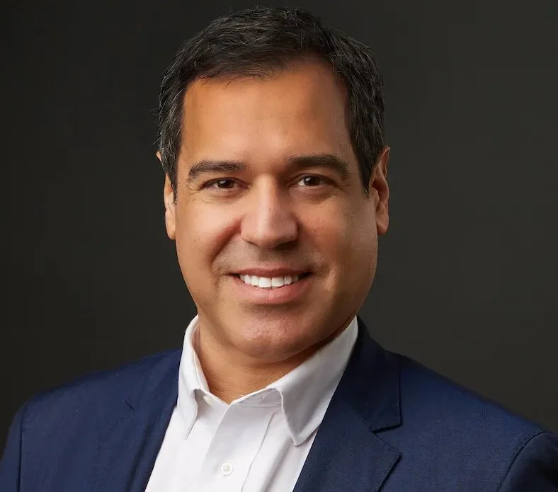 Portrait of a man with dark hair, wearing a white shirt and navy blazer, smiling against a dark background.