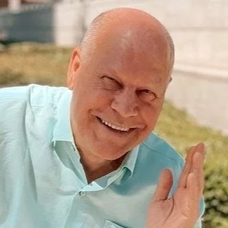A bald man smiling and waving outdoors