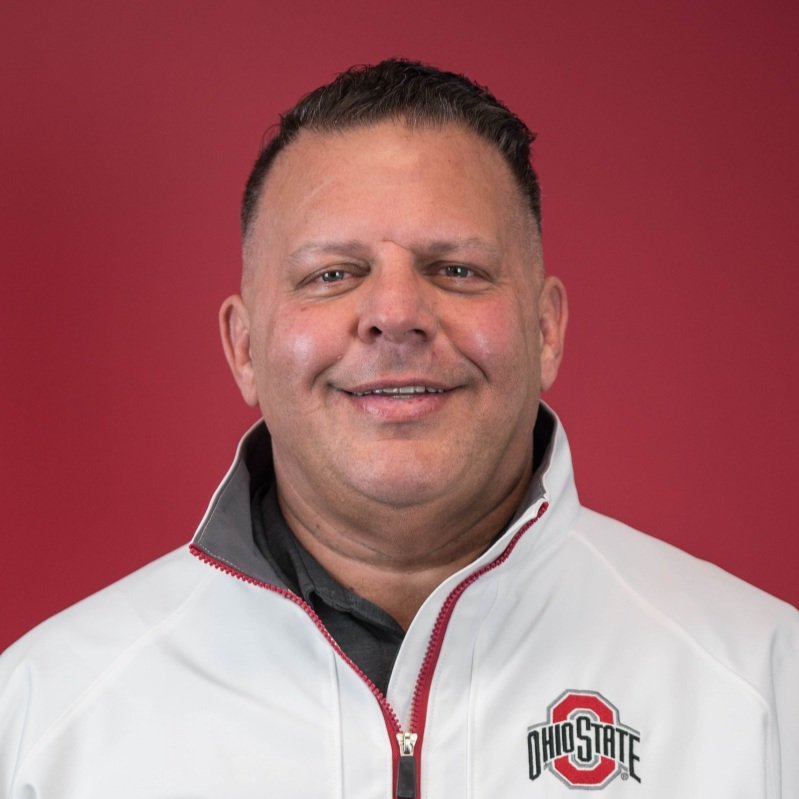A man with short dark hair smiling, wearing a white Ohio State jacket with a red background.