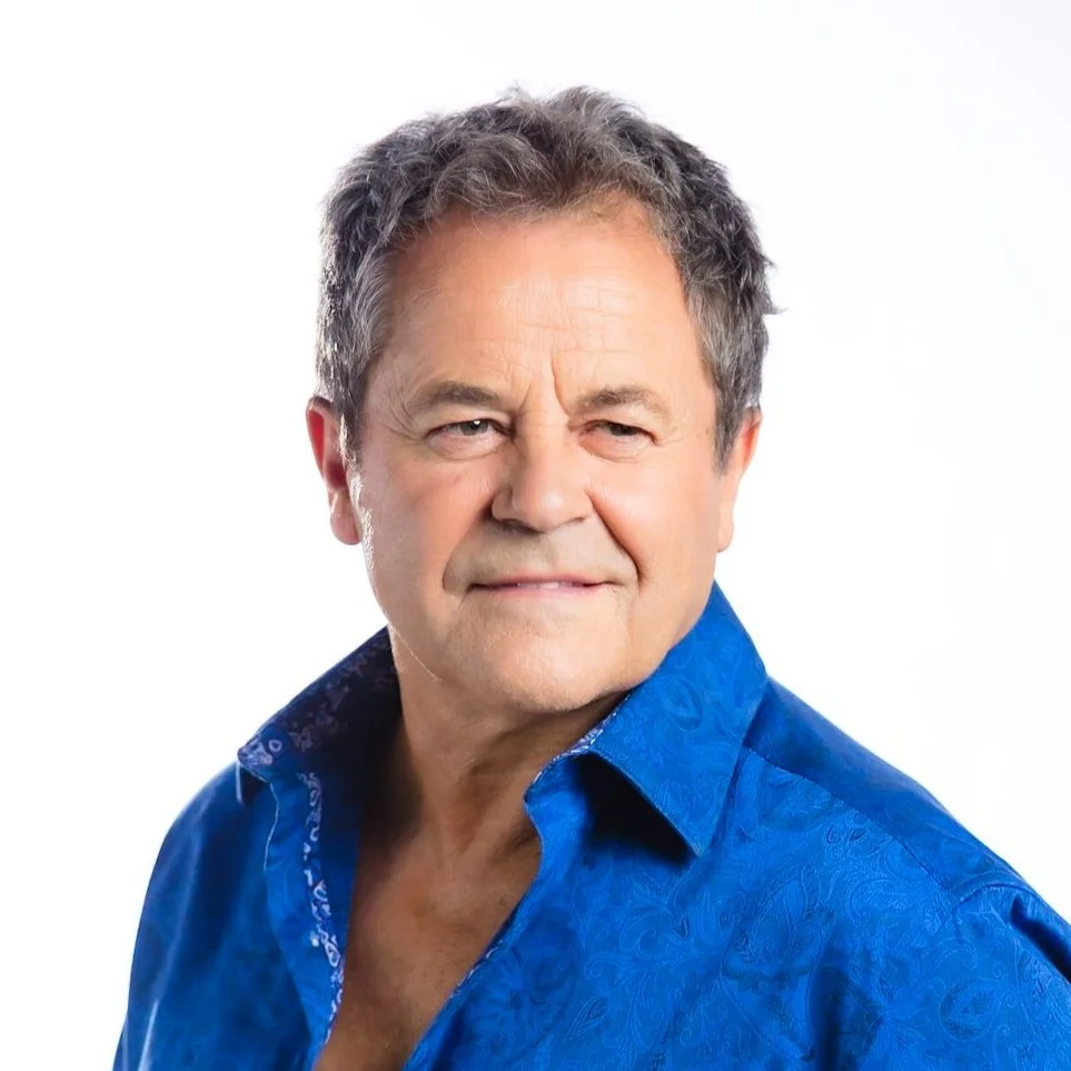 A middle-aged man with gray hair and a slight smile wearing a blue patterned button-up shirt against a plain white background.