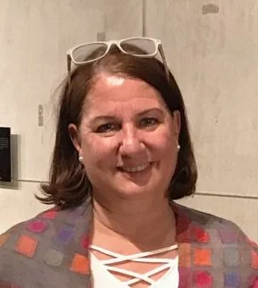 A smiling woman with shoulder-length brown hair wearing glasses on her head, a patterned blazer, and a white top with a crisscross design in the front, standing indoors against a neutral background.