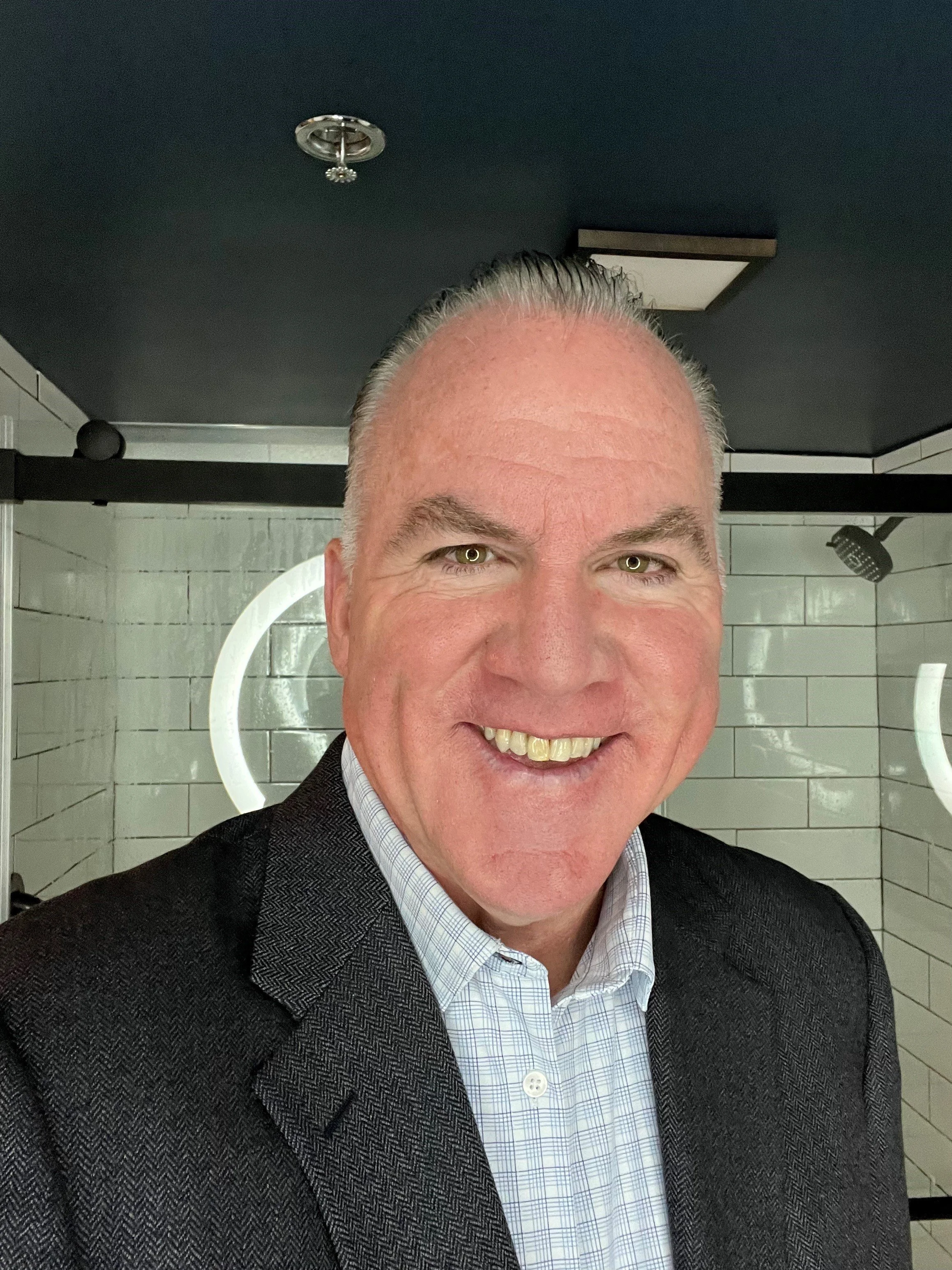 A smiling man with gray hair, wearing a dark suit and light checkered shirt, in a bathroom or dressing room with white tiled walls and black ceiling, illuminated by vanity mirror lights.