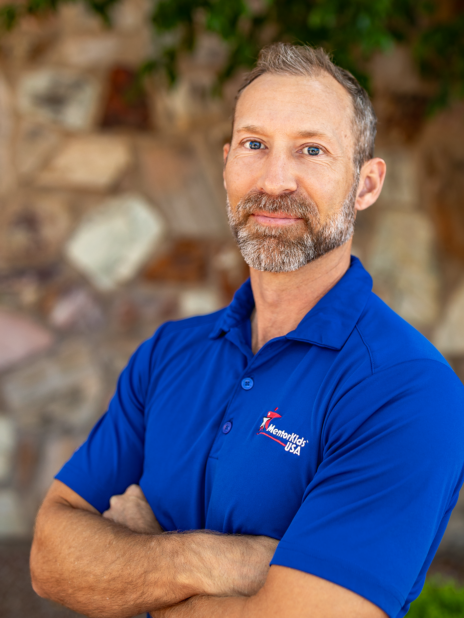 A middle-aged man with short hair and a beard wearing a blue polo shirt with a logo for Mentoring USA, standing outdoors with a stone wall and greenery in the background.