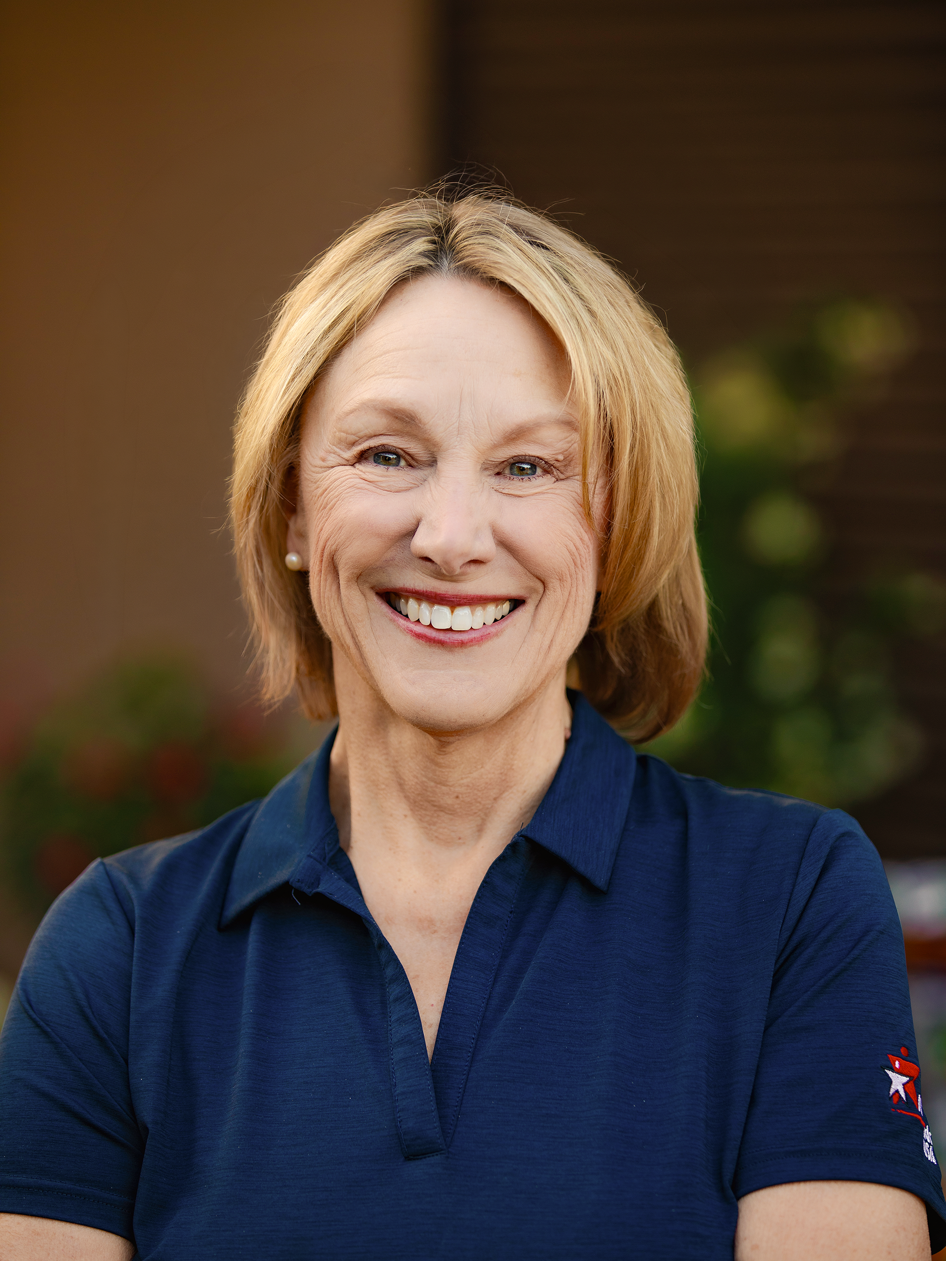 A smiling woman with blonde hair wearing a navy blue shirt with a patriotic star patch on the sleeve, outdoors with blurred greenery and building in the background.